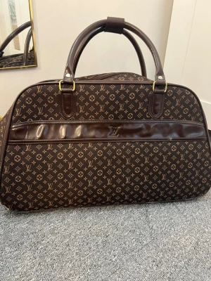 Brun Louis Vuitton resväska monogram - Stilren resväska från Louis Vuitton i klassiskt brunt monogrammönster. Väskan har två robusta handtag, guldfärgade metalldetaljer och dragkedja. Tillverkad i canvas med detaljer i läder. Perfekt för weekendresor och har en tidlös design.