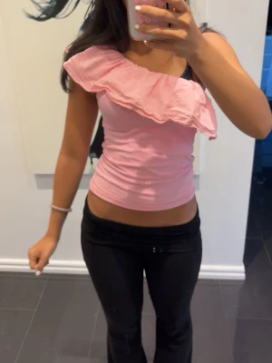 Rosa topp med volang - Säljer en söt rosa topp med omlott-design och stor volangdetalj över ena axeln. Toppen är ärmlös och har en tight passform som framhäver figuren. Perfekt för dig som vill sticka ut med en unik och trendig look.