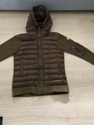 Brun quiltad cardigan med huva - Snygg brun cardiganjacka med quiltad framsida och stickade ärmar samt rygg. Jackan har huva, dragkedja framtill och fickor med knappstängning. Detalj med ficka på ena ärmen och ribbade muddar vid ärmslut och nederkant.