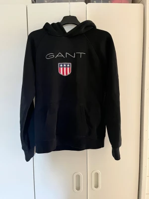 Svart hoodie från GANT med logga - Säljer en svart hoodie från GANT med stor vit logga och ett broderat rött, vitt och blått sköldmärke på bröstet. Hoodien har huva, känguruficka och ribbade muddar vid ärmar och nederkant. Perfekt för en avslappnad och stilren look. Storlek 158/164