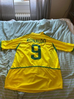 Gul Brasil Ronaldo 9 fotbollströja - Säljer en klassisk gul Brasilien fotbollströja med gröna detaljer och nummer 9 samt namnet Ronaldo på ryggen. Tröjan har broderad CBF-logga på bröstet och är tillverkad i ett lätt och ventilerande material, perfekt för fotboll eller samling.