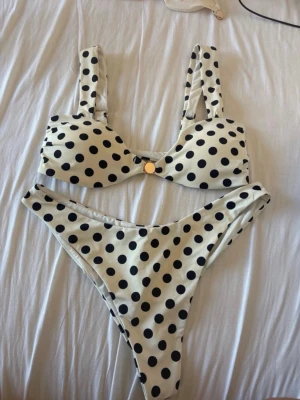 Vit bikini med svarta prickar - Snygg bikini i vitt med stora svarta prickar. Överdelen har breda axelband och en liten guldfärgad detalj framtill. Underdelen är högt skuren för en trendig look. Perfekt för stranden eller poolen! Överdel storlek S. Underdel finns i M och L