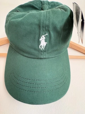 Grön keps från Ralph Lauren - Snygg grön keps från Ralph Lauren med klassisk vit broderad logga framtill. Kepsen är i bomullstyg och har böjd skärm. Perfekt accessoar för en avslappnad och trendig stil.