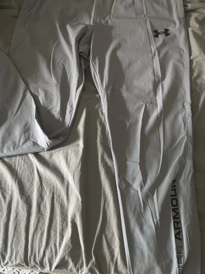 Grå träningsbyxor från Under Armour - Snygga grå träningsbyxor från Under Armour med logga på låret och text längs benet. Tillverkade i ett stretchigt och lätt material som passar perfekt för gymmet eller löpning. Resår i midjan för skön passform.