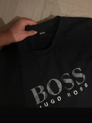 Svart sweatshirt från Hugo Boss - Svart sweatshirt från Hugo Boss med stort BOSS-tryck i vitt och silver på bröstet. Tröjan har rund halsringning och långa ärmar. Perfekt för en stilren och avslappnad look.