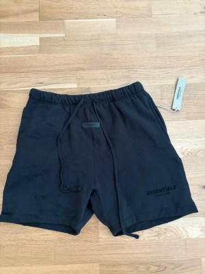 Essentials shorts  - Säljer ett par svarta shorts från Essentials Fear of God med broderad logga på benet och snörning i midjan. De har en avslappnad passform och är perfekta för sommaren. Tillverkade i mjuk bomull för maximal komfort.