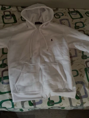 Vit zip hoodie med huva - Säljer en vit hoodie med dragkedja och huva. Tröjan har fickor framtill och justerbara snören i huvan. Klassisk och enkel design med liten broderad logga på bröstet. Perfekt för en avslappnad stil.