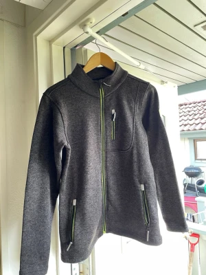 Mörkgrå fleecejacka med dragkedjor - Mysig mörkgrå fleecejacka med hel dragkedja framtill och kontrasterande gröna detaljer på fickor och bröstficka. Jackan har hög krage, två sidofickor och en bröstficka med dragkedja. Perfekt för kyliga dagar och enkel att matcha med olika outfits.
