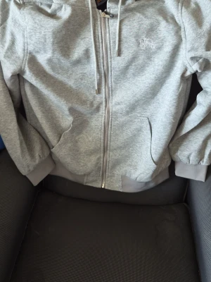 Ljusgrå hoodie med dragkedja - Snygg ljusgrå hoodie med dragkedja framtill och två fickor. Tröjan har huva med dragsko och ribbade muddar vid ärmar och nederkant. Materialet är mjukt och bekvämt, perfekt för chill dagar. Liten broderad logga på bröstet.