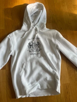 Vit hoodie med tryck från BVALDI - Snygg vit hoodie från BVALDI med svart tryck framtill som föreställer ett stilrent emblem och text. Hoodien har klassisk känguruficka, dragsko i huvan och mjukt material. Perfekt för en avslappnad och trendig look.