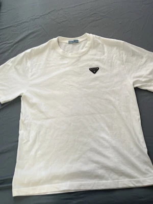 Vit t-shirt med Prada logga på bröstet - Klassisk vit t-shirt i bomull med rund hals och korta ärmar. På vänstra bröstet sitter en liten svart logga som ger en stilren touch. Perfekt basplagg för alla tillfällen och enkel att matcha med andra plagg. Det är storlek M men passar som L. Pris kan diskuteras vid snabb affär. 