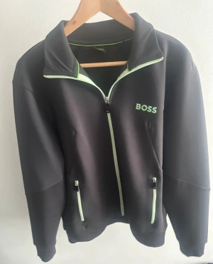 Boss kofta - Snygg grå ziptröja från BOSS med limegröna detaljer längs dragkedjor och krage. Tröjan har hög krage, två fickor med dragkedja och BOSS-logga broderad på bröstet. Perfekt för en sportig och stilren look.