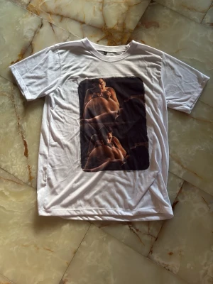 Vit t-shirt med tryck - Vit t-shirt med korta ärmar och ett stort tryck framtill som visar ett konstnärligt foto i mörka färger. T-shirten har en klassisk passform och rund halsringning. Perfekt för dig som vill ha en unik och uttrycksfull stil. (Grish)