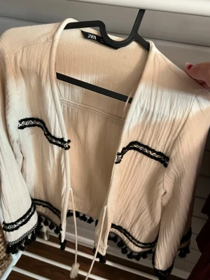 Beige bolerotopp med svarta detaljer - Säljer en beige bolerotopp från ZARA med svarta broderade band och tofsar längs kanterna. Toppen har knytband framtill och långa ärmar. Perfekt för att lyfta en enkel outfit med lite boho-vibe.