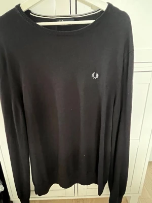 Svart stickad tröja från Fred Perry - Klassisk svart stickad tröja från Fred Perry med rund halsringning och diskret logga broderad på bröstet. Tröjan har långa ärmar och ribbade muddar vid ärmslut och nederkant. Perfekt för en stilren och enkel look. 54% ull. Storlek XL
