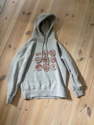 Grå hoodie Maison Kitsuné Hoodie - Snygg grå hoodie från Maison Kitsuné med brunt tryck framtill som visar olika emblem och loggor. Hoodien har huva med dragsko och ribbade muddar vid ärmar och midja. Perfekt för en chill och trendig look. Helt ny, med tags.