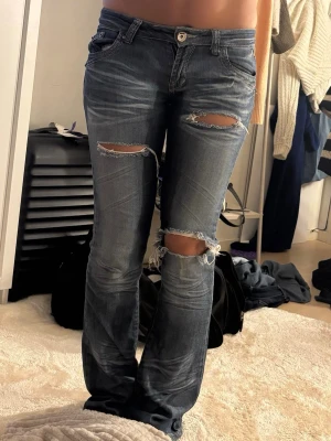 Blå vintage bootcut jeans - Säljer ett par blå bootcut jeans med slitningar och stora hål på låren och knäna. Jeansen har klassisk femficksdesign, låg midja och dragkedja med knapp. Perfekta för en avslappnad och trendig look. Dragkedjan är lite trög men fungerar att dra upp och ner som den ska!