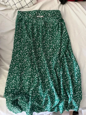 Grön blommig långkjol med resårmidja - Säljer en grön långkjol med små vita blommor och resår i midjan. Kjolen har en luftig och flowy passform, perfekt för varma dagar. Tyget känns mjukt och lätt, vilket gör den superbekväm att bära.