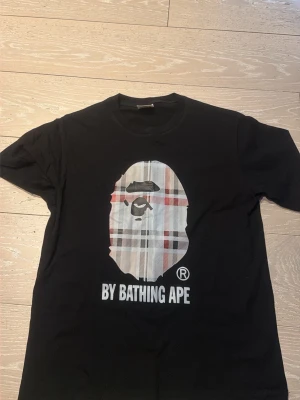 Svart BAPE t-shirt med rutig logga - Säljer en svart t-shirt från BAPE med den ikoniska apansiluetten i rutigt mönster på bröstet och texten 'BY BATHING APE' under. Klassisk passform och rund hals. Perfekt för dig som gillar streetwear och vill sticka ut.