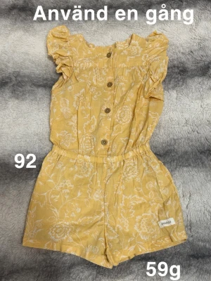 Gul blommig jumpsuit med volangärm - Söt gul jumpsuit med vita blommönster och volangärmar. Knappar framtill och resår i midjan för skön passform. Kortbent modell i lätt och mjukt tyg, perfekt för varma dagar. Liten etikett på benet.🌸 Använd en gång. Storlek 92