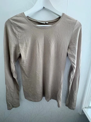 Beige långärmad topp från Kappahl - En enkel och stilren beige långärmad topp från Kappahl i mjuk bomull. Modellen har rund halsringning och en normal passform som funkar till många olika looks. Perfekt basplagg att ha i garderoben.
