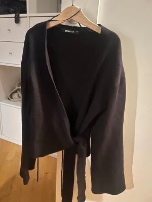 Svart omlottkofta från Gina Tricot - Stilren svart omlottkofta från Gina Tricot med knytband i midjan och långa ärmar. Koftan har en mjuk och tunn stickad känsla, perfekt att slänga över en topp eller klänning. Enkel att styla och passar till många olika looks.