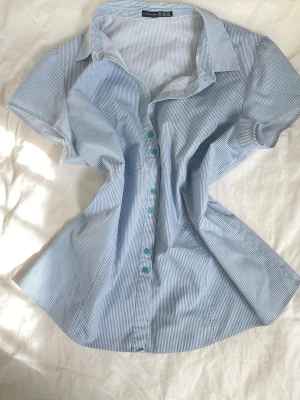 Blårandig kortärmad blus - Snygg blus med tunna blå och vita ränder, klassisk krage och blå knappar framtill. Modellen är figursydd och har korta ärmar. Perfekt till jeans eller kjol för en fräsch look. Materialet känns lätt och luftigt.