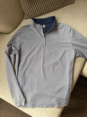 FootJoy Golf Pullover 1/4 Zip – Herr L - Säljer en stilren FootJoy golftröja med 1/4 dragkedja i storlek L. Tillverkad i ett lätt och stretchigt funktionsmaterial som passar perfekt både på golfbanan och till vardags. Den grå färgen tillsammans med marinblå detaljer ger ett klassiskt och tidlöst utseende.  Tröjan är i mycket fint skick utan anmärkningar och är perfekt som ett extra lager under kyligare rundor eller som en tunn vardagströja.