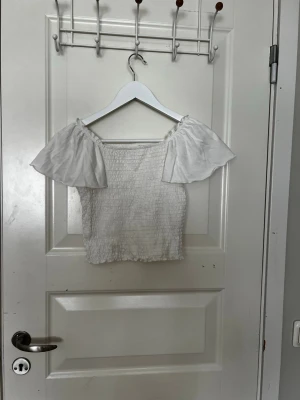 Vit smockad topp från Kappahl - Supersöt vit topp från Kappahl med smockad kropp och vida, korta offshoulder-ärmar. Perfekt till sommaren och lätt att matcha med både jeans och kjol. Luftig och bekväm.