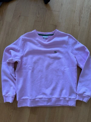 Rosa sweatshirt från Lacoste - Säljer en rosa sweatshirt från Lacoste med klassisk rund halsringning och ribbade muddar. storlek 164,14 år. Tröjan har den ikoniska gröna krokodilloggan broderad på bröstet. Tillverkad i mjukt bomullsmaterial för en skön känsla.