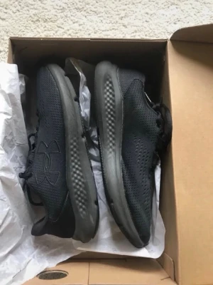 Svarta Under Armour sneakers - Snygga helsvarta sneakers från Under Armour med mesh-ovandel för bra ventilation och en tjock, dämpad sula. Skorna har en låg profil och diskret logga på sidan. Perfekta för dig som gillar stilrena och sportiga skor. Använd 2 gånger