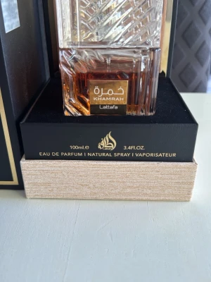 Lattafa Khamrah Eau de Parfum 100ml - Lattafa Khamrah Eau de Parfum i en elegant, fyrkantig glasflaska med detaljerat mönster och guldiga inslag. Flaskan rymmer 100 ml och kommer i en lyxig svart och guld förpackning med trädetalj. Cirka 87-90ml kvar