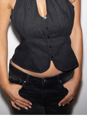 Darling denim topp - Snygg svart korsettopp i denim-look med halterneck och knytband i nacken. Toppen har knappar framtill och markerade sömmar som ger en cool och edgy vibe. Den är i storlek S