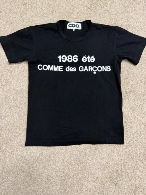 Svart Comme des Garçons t-shirt - Säljer en svart t-shirt från Comme des Garçons med vit text '1986 été COMME des GARÇONS' på bröstet. 