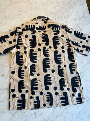 Beige skjorta med svart abstrakt mönster, NN07 - Kortärmad skjorta i beige med svart abstrakt mönster och knappar framtill. Skjortan har en avslappnad passform och klassisk krage. Perfekt för dig som gillar unika prints och vill sticka ut lite extra. Skjortan är av modell bowlingskjorta. Oanvänd. Nypris 1300 SEK. 