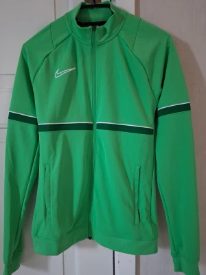 Grön träningströja från Nike - Snygg grön träningströja från Nike med hel dragkedja framtill och vita detaljer. Tröjan har två fickor, lång ärm och en vit Nike-logga på bröstet. Svarta och vita ränder går tvärs över bröstet och armarna för en sportig look.