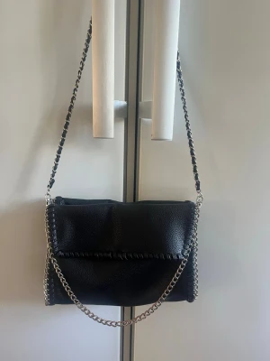 Svart axelväska med kedja Gina Tricot - Snygg svart axelväska från Gina Tricot i fejkläder med silverfärgad kedjerem och dekorativa sömmar längs kanterna. Väskan har ett innerfack med dragkedja och är perfekt för att bära det viktigaste. Enkel men trendig design som passar till många outfits.