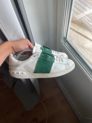 Valentino Open  - Snygga vita sneakers med en bred grön rem över mitten och vita snören. Skorna har en låg profil och detaljerade sömmar längs sidorna. Tillverkade i slätt läder med en klassisk sula och diskreta nitar vid hälen.
