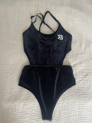 Svart sammetsbody från Better Bodies  - Stilren svart body i sammet med smala axelband som korsas i ryggen. Hög benskärning och en vit logga framtill. Med cut-outs på sidan.  Perfekt att styla till jeans eller kjol för en trendig look.
