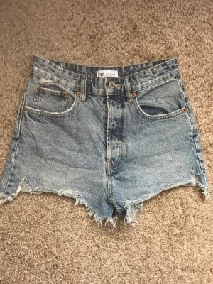 Blå jeansshorts med rå kant från ZARA - Snygga blå jeansshorts från ZARA med hög midja och rå, fransig kant. Storlek är 36 men funkar även för storlek 34. Knappgylf och sliten urtvättad look. Klassisk femficksmodell i slitstarkt denimtyg. Perfekta för en avslappnad och trendig sommarlook. 