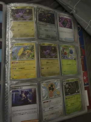 Pokémonkort i samlarpärm - Säljer en sida med Pokémonkort i en genomskinlig samlarpärm. Korten har olika motiv och färger, bland annat lila, gul, grön och metallisk. Texten på korten är på koreanska och de har olika illustrationer av Pokémon och tränare. Perfekt för dig som gillar att samla Pokémonkort.