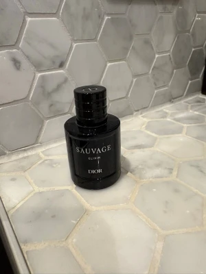 Sauvage Elixir parfym från Dior - Sauvage Elixir från Dior är en exklusiv herrparfym i en mörkblå, nästan svart, cylinderformad glasflaska med elegant silvertext. Flaskan har en stilren och lyxig design som känns modern och tidlös. Volymen är 7,5 ml, perfekt att ta med sig.