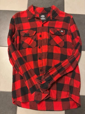 Röd och svart rutig skjorta Dickies - Klassisk röd och svart rutig skjorta från Dickies i bomull. Skjortan har två bröstfickor med lock och knappar, samt en avslappnad passform och långa ärmar. Perfekt för en cool och avslappnad stil.