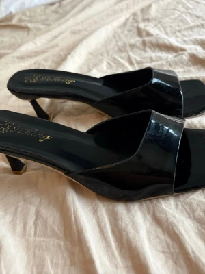 Svarta lackade slip-in pumps - Säljer ett par svarta slip-in pumps med öppen tå och fyrkantig framdel. Skorna har en låg klack och är tillverkade i glansigt lackmaterial. Perfekta för dig som vill ha en stilren och trendig look. väldigt enkla att gå med 