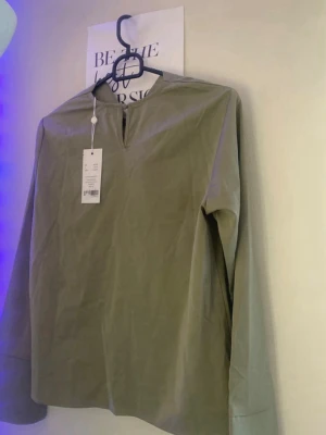 Olivgrön långärmad blus - Stilren olivgrön blus med lång ärm och enkel V-ringning utan knappar. Blusen har en rak passform och är tillverkad i ett mjukt, tunt tyg som ger en avslappnad look. Perfekt att matcha med jeans eller kjol för en trendig vardagsstil.
