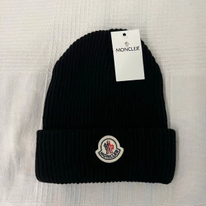 Svart ribbad mössa från Moncler - Svart ribbstickad mössa från Moncler med uppvikt kant och klassisk Moncler-logga framtill. Mössan har en enkel och stilren design som passar till det mesta. Perfekt accessoar för kalla dagar. Kontakta vid frågor eller funderingar😊