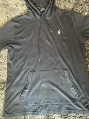 Mörkblå hoodie från Polo Ralph Lauren - Snygg mörkblå hoodie från Polo Ralph Lauren med klassisk huva och dragsnören. Har en stor magficka och den ikoniska vita Polo-loggan broderad på bröstet. Tillverkad i mjukt bomullsmaterial som känns skönt mot huden. Perfekt för en avslappnad och stilren look.
