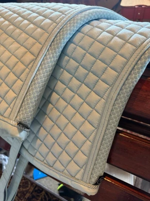 Ljusblått quiltat schabrak Eskadron - Säljer ett ljusblått schabrak med quiltat rutmönster och snygga detaljer längs kanterna. Perfekt till ridning och ger en stilren look till din hästutrustning. Schabraket har praktiska fästen och är tillverkat i ett mjukt material. Säljes tvättat, i fint skick men med lite hår kvar och några mindre fläckar som syns på bild.