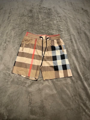 Burberry badbyxor - Snygga shorts i beige med klassiskt rutigt mönster i svart, vitt och rött. Shortsen har elastisk midja med svart snörning och sidofickor. Perfekta för sommaren och har en avslappnad passform.