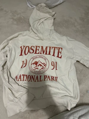 Kappahl  - Vit hoodie med röd text och tryck framtill där det står 'Yosemite National Park 1991' och ett bergmotiv. Hoodien har huva och ribbade muddar vid ärmslut och nederkant. Perfekt för dig som gillar avslappnad och cool stil.